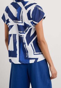 Camicia a maniche corte in tessuto bianco e blu, caratterizzata da motivi geometrici astratti. Vestibilità comoda con colletto e maniche leggermente arrotolate.