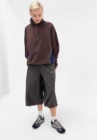 Brązowa bluza na suwak z niebieskimi panelami po bokach, zestawiona z kraciastymi, skróconymi spodniami oraz czarnymi butami sportowymi z fioletowymi akcentami.