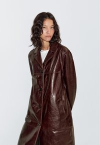 Bruine leren jas met een getailleerd ontwerp, voorzien van een gekerfde kraag, zes knopen en zijzakken. Gladde textuur en knielengte.