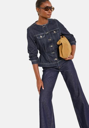 Žena v tmavé džínové bundě a zvonových džínách drží karamelovou semišovou kabelku, má sluneční brýle a zlaté kruhové náušnice.