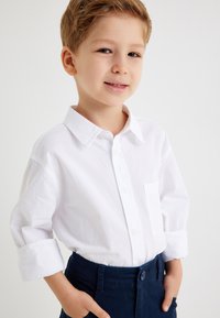 Biała koszula z długim rękawem, zapinana na guziki, z małą kieszonką, połączona z granatowymi spodniami. Materiał wydaje się gładki i lekki.