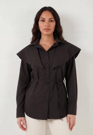 Kvinde med bølget mørkt hår iført sort skjorte med knapper, bred krave og bælte, kombineret med lyse bukser, står foran en ensfarvet baggrund.