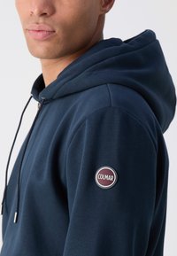 Felpa con cappuccio blu navy realizzata in morbido cotone, con chiusura a zip e un patch rotondo del logo Colmar sulla manica sinistra.