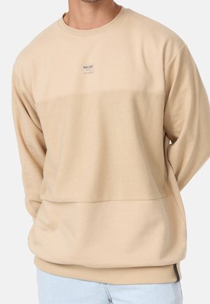 Mann trägt beige langärmeligen Sweatshirt mit strukturiertem Einsatz und kleinem "INDICODE" Logo-Patch auf der Brust, kombiniert mit hellblauen Jeans.