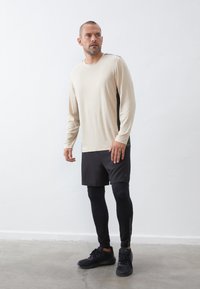 Lichtbeige lange mouw top met zwarte zijpanelen, gecombineerd met zwarte shorts en leggings. De outfit heeft een gladde textuur en een sportieve pasvorm.