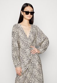 Vestido de estampa de leopardo em tecido bege claro, apresentando um decote em V profundo, mangas longas e volumosas, e detalhes drapeados. Óculos de sol retangulares pretos incluídos.