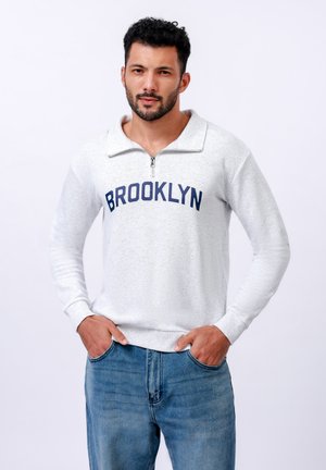 Svetlo siva majica sa četvrtinastim patentnim zatvaračem, sa natpisom "BROOKLYN" u mornarsko plavoj boji preko grudi. Tekstura tkanine, duži rukavi i rebrasti manžeti.