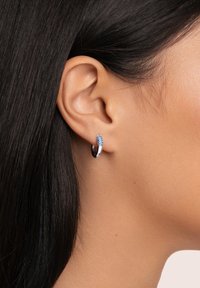 Orecchino a cerchio argentato con accentuazione cristallizzata blu, superficie liscia e forma circolare. Indossato in un primo piano di un orecchio.