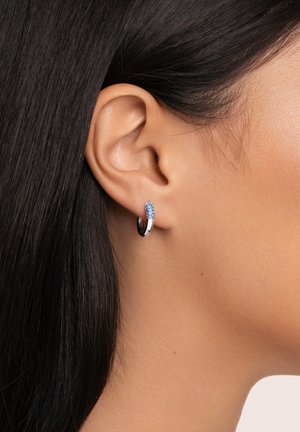 Orecchino a cerchio argentato con accentuazione cristallizzata blu, superficie liscia e forma circolare. Indossato in un primo piano di un orecchio.