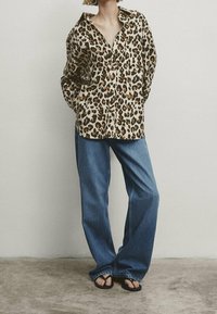 Chemise à boutons imprimé léopard en tissu léger, associée à un jean denim bleu ample et des sandales noires, présentée sur un fond neutre.