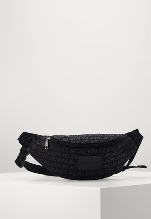 Schwarze Bauchtasche von Calvin Klein Jeans mit Reißverschluss-Hauptfach und verstellbarem Riemen, auf einer weißen Oberfläche platziert.
