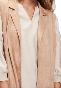 Beżowa zamszowa kamizelka z otwartym przodem, dekoltem z wycięciem i dwoma kieszeniami po bokach. Pod spodem jasnobeżowa koszula z dekoltem w serek i elastycznymi mankietami.