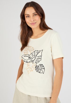 Mujer con cabello largo y castaño que viste una camiseta crema con ilustración lineal de hojas en negro y naranja y pantalones beige a rayas, de pie frente a un fondo gris.