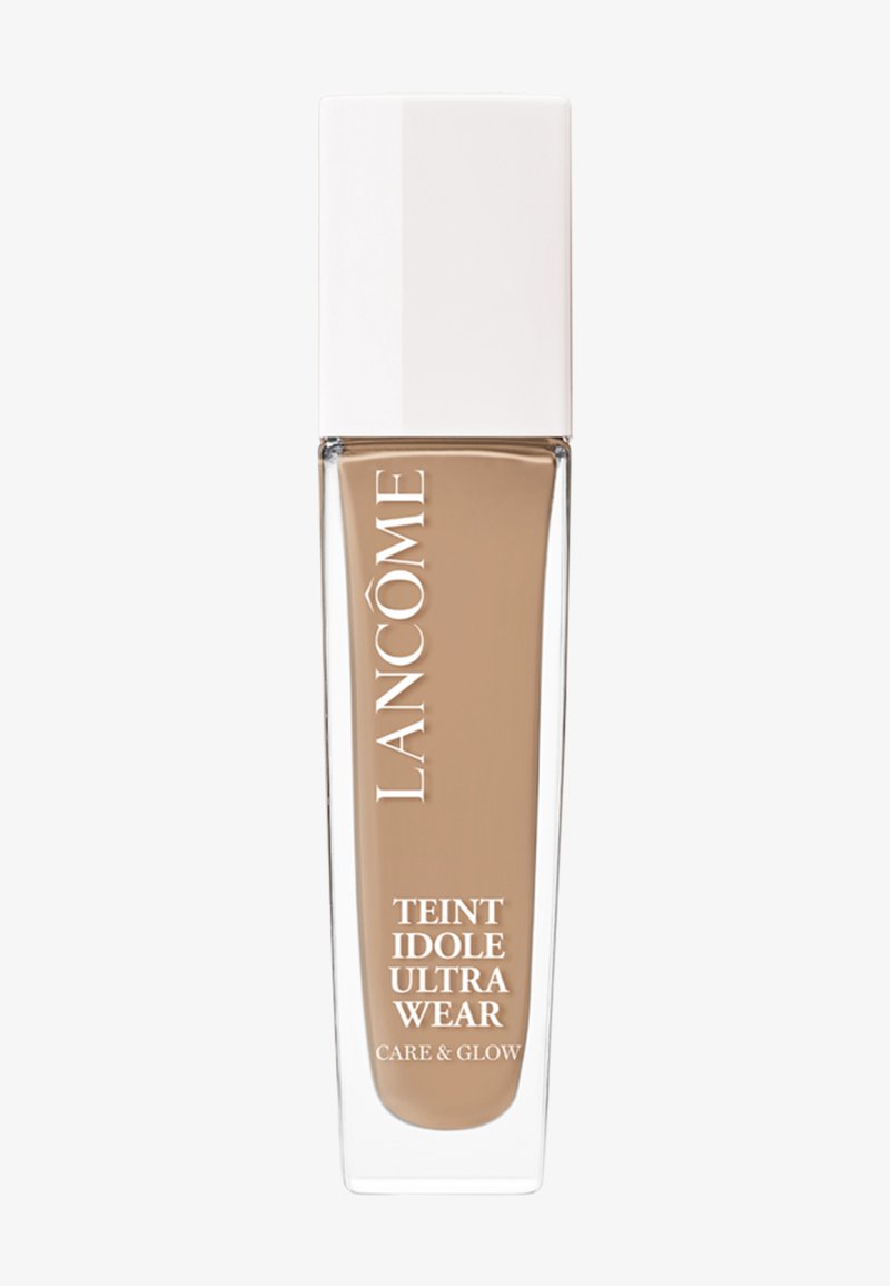 Fond de teint dans une bouteille en verre transparent de forme rectangulaire, avec un bouchon carré blanc. Le liquide est d'une teinte beige clair.