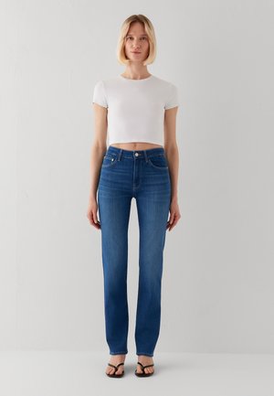 Frau mit kurzem blondem Haar, die ein weißes bauchfreies T-Shirt, blaue hoch taillierte Jeans und schwarze Sandalen trägt, steht vor einer schlichten grauen Wand.