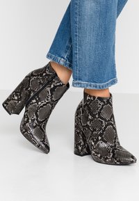 Piedi che indossano stivali ankle con tacco alto dal motivo in serpente nero e grigio, abbinati a jeans di denim blu a vita corta su un pavimento bianco.