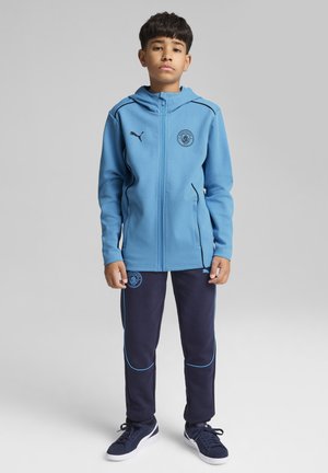 Haut à capuche zippé bleu clair avec des bordures noires et un emblème, associé à un pantalon bleu foncé avec des accents bleus, complété par des chaussures sombres.