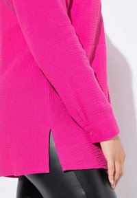 Fuchsia Langarmshirt aus strukturiertem Stoff, mit seitlichen Schlitzen und geknöpften Manschetten, kombiniert mit schwarzen Leggings.