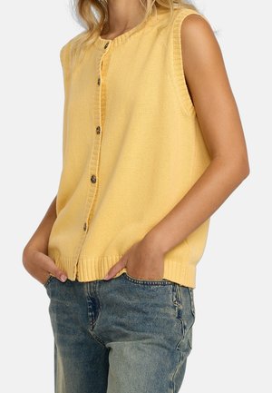 Person trägt ärmellose gelbe Strickweste mit Knöpfen und blaue Jeans, Hände in den Taschen, vor einem einfarbigen Hintergrund.