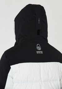 Svart och vit puffjacka med huva, texturerat tyg och "GORE-TEX INFINIUM"-logga på baksidan.