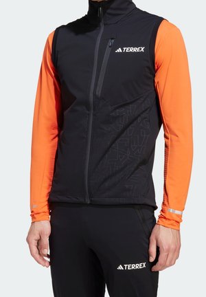 Man draagt een zwart vest met rits en TERREX-logo over een feloranje shirt met lange mouwen en zwarte broek met bijpassend logo, staat met ontspannen armen.