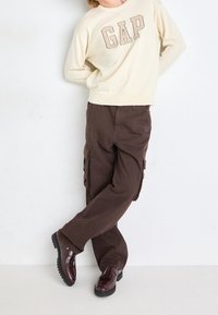 Krämfärgad tröja med "GAP"-logotyp, bruna cargobyxor och vinröda skor med en texturerad sula. Outfitten har avslappnad passform och en avslappnad stil.