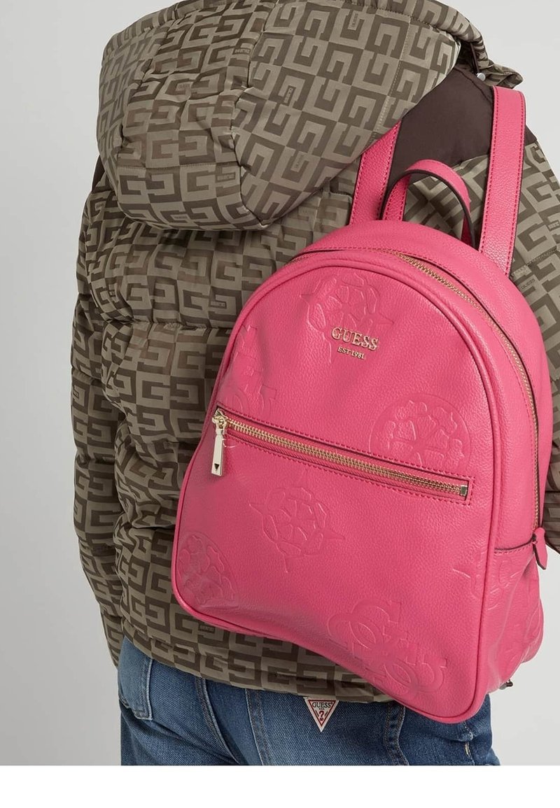 Personne portant une veste marron à motifs et un jean bleu avec un sac à dos Guess rose vif avec une fermeture éclair dorée et des logos en relief.