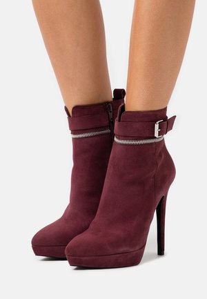 Bottines en suède bordeaux avec un bout pointu, un haut talon aiguille, une bride à la cheville et un détail de fermeture éclair en métal décoratif.