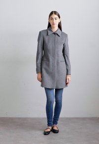 Woman standing against gray wall wearing gray knee-length coat, blue skinny jeans, and black ballet flats, facing forward with neutral expression.