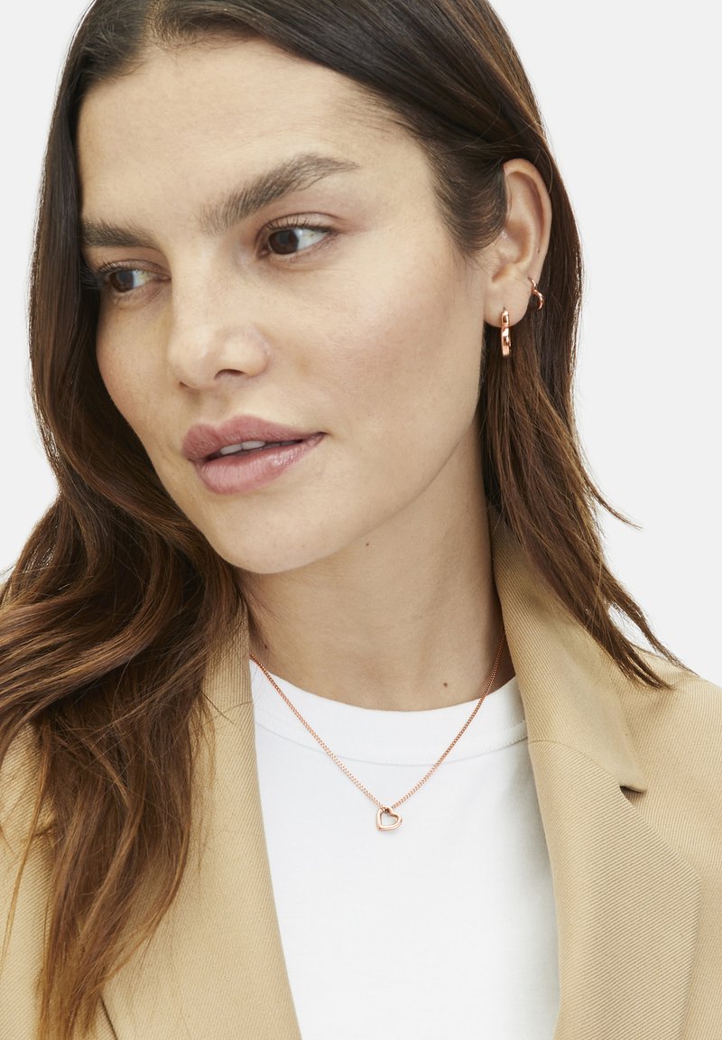 Gold-tone jewelry features a delicate heart pendant on a thin chain, with matching hoop earrings, worn with a beige blazer and white top.