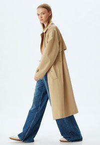 Beige trenchcoat med vida ärmar och knappar, parat med högmidjade blå denim jeans med vida ben och vita sneakers.