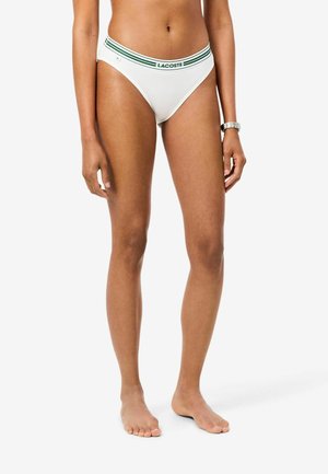 Femme portant un bas de bikini blanc Lacoste avec des rayures vertes et un logo sur la ceinture, debout pieds nus sur un fond uni.