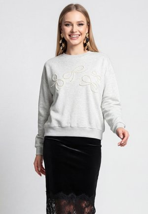 Smilende kvinde iført store guldbøjler, lysegrå sweater med perlebuer og sort fløjlsnederdel med blondekant, stående på grå baggrund.
