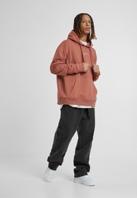 Rustfarvet hættetrøje med en kengurulomme, parret med løse sorte bukser og hvide sneakers, sat mod en neutral baggrund.