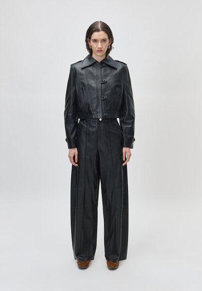 Žena stoji na jednobojnoj pozadini, nosi crnu kožnu jaknu do struka i široke kožne hlače visokog struka s smeđim cipelama.
