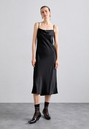 Black satin slip dress with thin straps, cowl neckline, and a flowing hem. Paired with black sheer socks and high-heeled shoes.