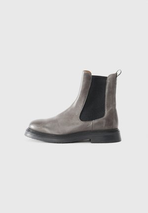 Bottine en cuir gris avec des panneaux élastiques sur les côtés, semelle noire plate et une petite poignée. Texture lisse avec un design à bout arrondi.