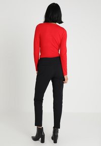 Vrouw met zwart haar, gekleed in een rode blouse met lange mouwen, zwarte slim-fit broek en zwarte enkellaarsjes, staand met de rug naar de camera op een witte achtergrond.