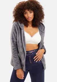 Veste grise, texturée et duveteuse avec une capuche, dotée d'une fermeture éclair à l'avant et de poches latérales, portée par-dessus un bralette blanc à dentelle et un pantalon bleu marine.