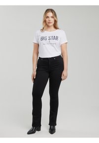 Naine kannab valget "BIG STAR" särk, musti kõrge vöökohaga teksaseid ja musti kontsakingi, seistes halli taustaga vastu.