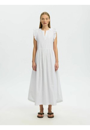 Femme aux cheveux raides portant une robe midi blanche sans manches avec des épaules à volants et des sandales noires à bouts ouverts, debout sur un fond blanc.