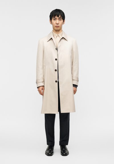 Trench-coat beige clair avec col, doté de boutons foncés. Porté sur une chemise claire et un pantalon à fines rayures, associé à des chaussures noires.