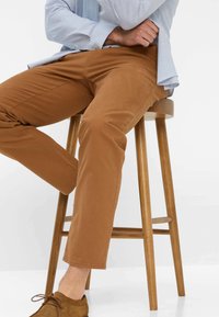 Pantaloni in cotone marrone, design a gamba dritta, con un leggero lucido, abbinati a una camicia a righe mentre si è seduti su uno sgabello di legno.