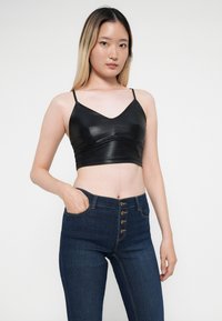 Fekete műbőr cropped top vékony pántokkal és V-nyakkivágással, amelyet magas derekú sötétkék farmernadrággal párosítanak, öt gombos részletekkel.