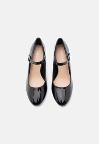 Zapatos Mary Jane de charol negro con punta redondeada, tira de tobillo y un interior suave. Nombre de la marca visible en la plantilla.