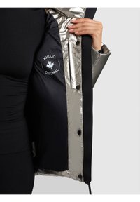 Persona con un outfit nero che tiene aperta una giacca argento e nera mostrando la tasca interna con il logo "Khujo Original" e bottoni a pressione.