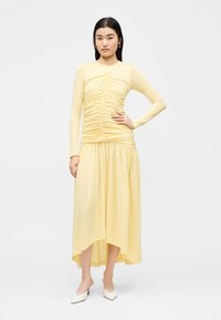 Γυναίκα στέκεται με το χέρι στη μέση, φορώντας μακρυμάνικο κίτρινο φόρεμα με πιέτες και λευκά μυτερά τακούνια, με μονόχρωμο φόντο.