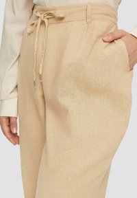 Beige broeken van een gestructureerde stof met een elastische tailleband en trekkoord, zijzakken en een relaxte pasvorm. Subtiele glans op het materiaal.