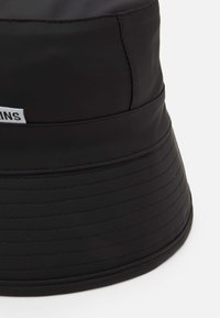 Black bucket hat made of textured fabric with a smooth finish. Features horizontal stitching and a small brand label on the side.