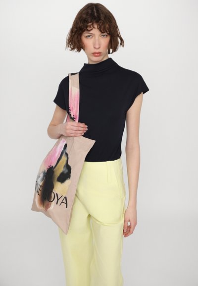 Sac en toile avec des motifs abstraits rose, noir et beige ; le texte indique "INE GOYA." Le modèle porte un haut noir et un pantalon jaune.